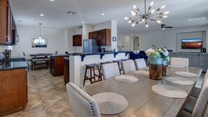 A modern kitchen with a central island, a dining area with a long table and chairs, and a living space with a sofa and a TV.