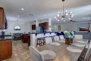 A modern kitchen with a central island, dining area with round tables, and a large chandelier. - Minutes to Coachella · Pool & Spa · 5BR Sleeps 12 (Indio)
