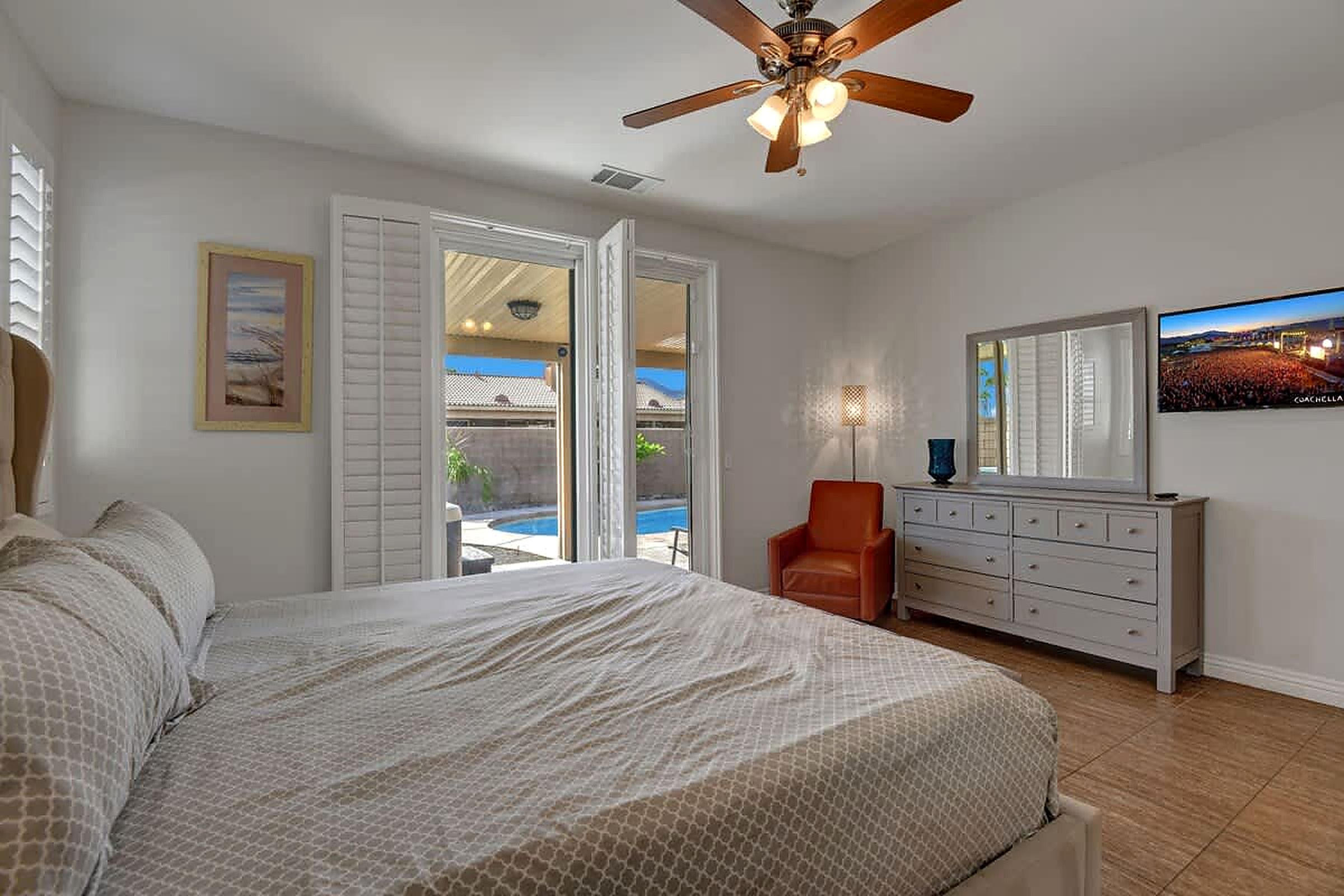 Un dormitorio con cama, cómoda, silla, ventilador de techo y ventana con vista a una piscina y edificios.