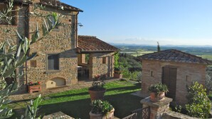 Un bâtiment en pierre avec un toit en tuiles, une cour agrémentée de plantes en pot et une vue sur des collines lointaines.