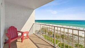 A balcony with a red Adirondack chair, a white railing, and a view of a beach with clear blue water and a sandy shore. - The Summit 727 (Panama City Beach)