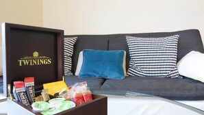 Ein Wohnzimmer mit grauem Sofa, einem Glastisch und einer braunen Schachtel mit dem Aufdruck "TWININGS", die verschiedene Lebensmittel enthält.