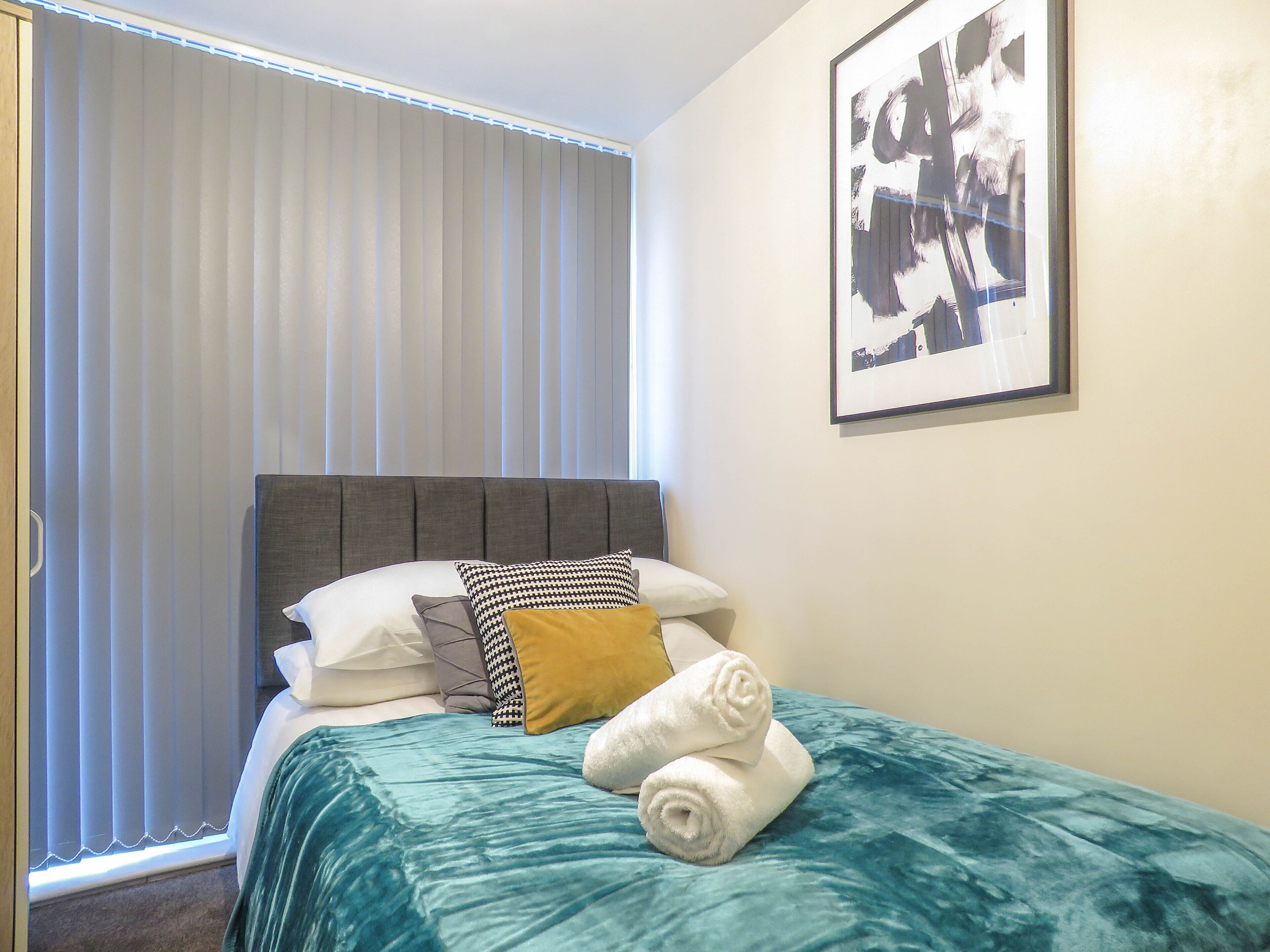 A hotel room with a bed, a window with blinds, a framed picture on the wall, and a door.