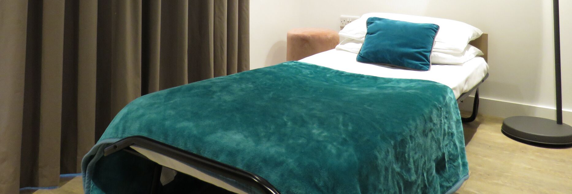 A room with a bed covered in a teal blanket, a white pillow, and a blue pillow. There is a brown ottoman beside the bed and a black floor lamp to the right.