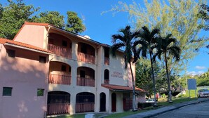 Un edificio de dos plantas con balcones, un letrero que dice "Residencia Nadiña Place", y vista de la calle con palmeras y coches estacionados.