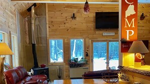 A cozy cabin interior with a wooden interior, a fireplace, a dining area, and a view of the snowy forest outside. - Rangeley Getaway (Dallas Plantation)