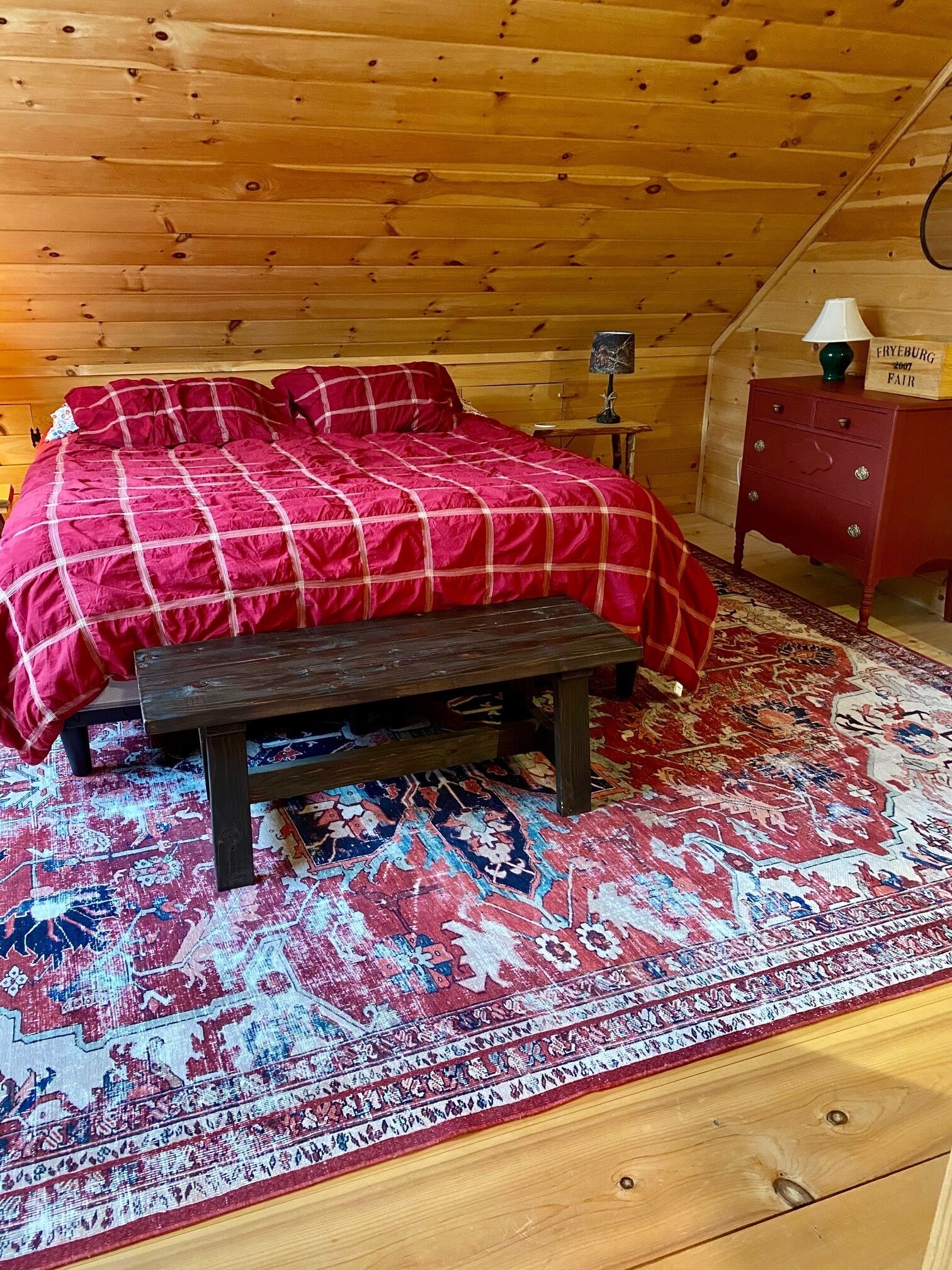 A cozy bedroom with a wooden ceiling, a bed with a red checkered bedspread, a bench, and a red dresser.