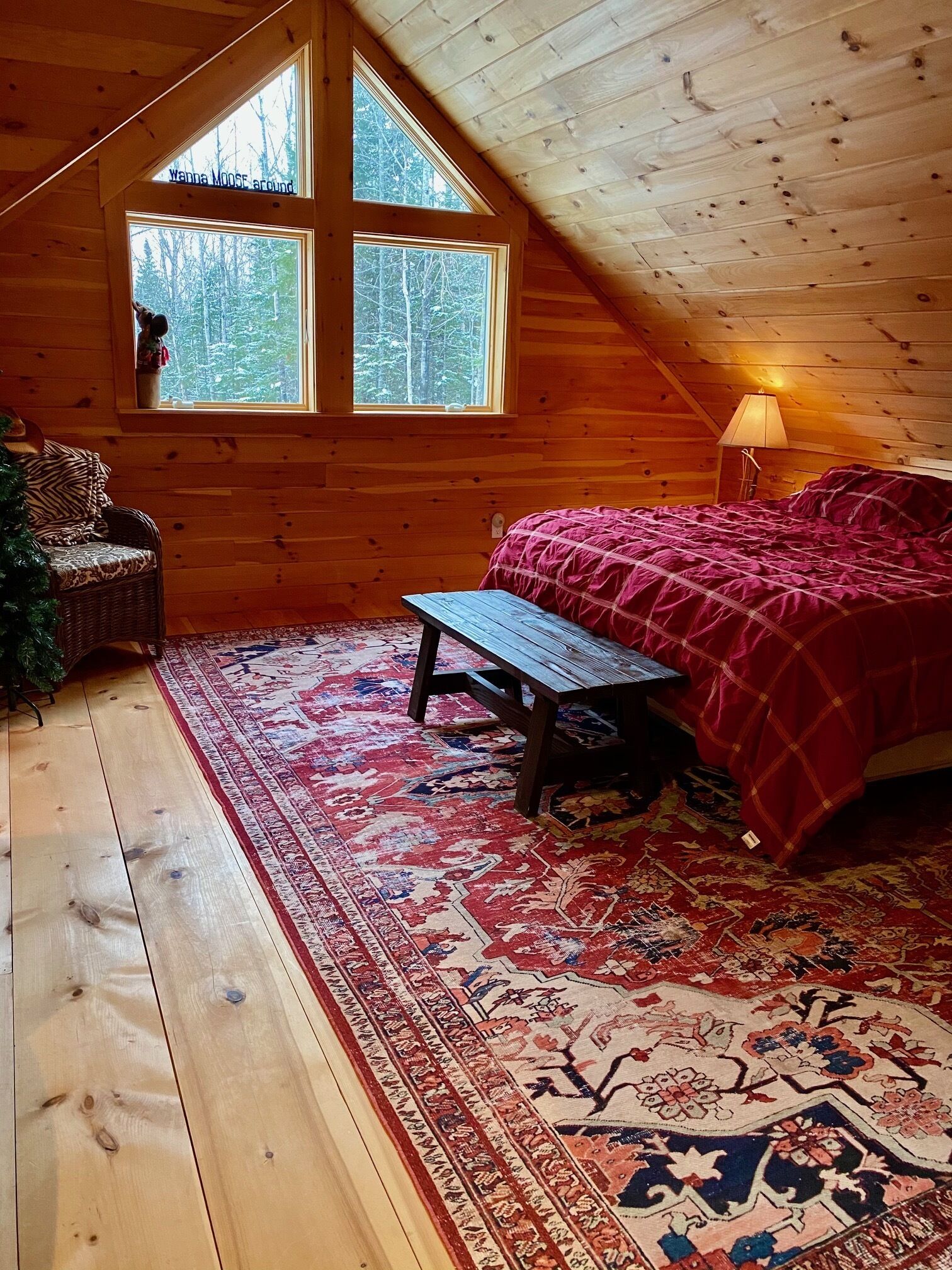 Camera da letto accogliente in un rifugio, con soffitto in legno, un letto grande con coperta rossa, una panca e un tappeto.