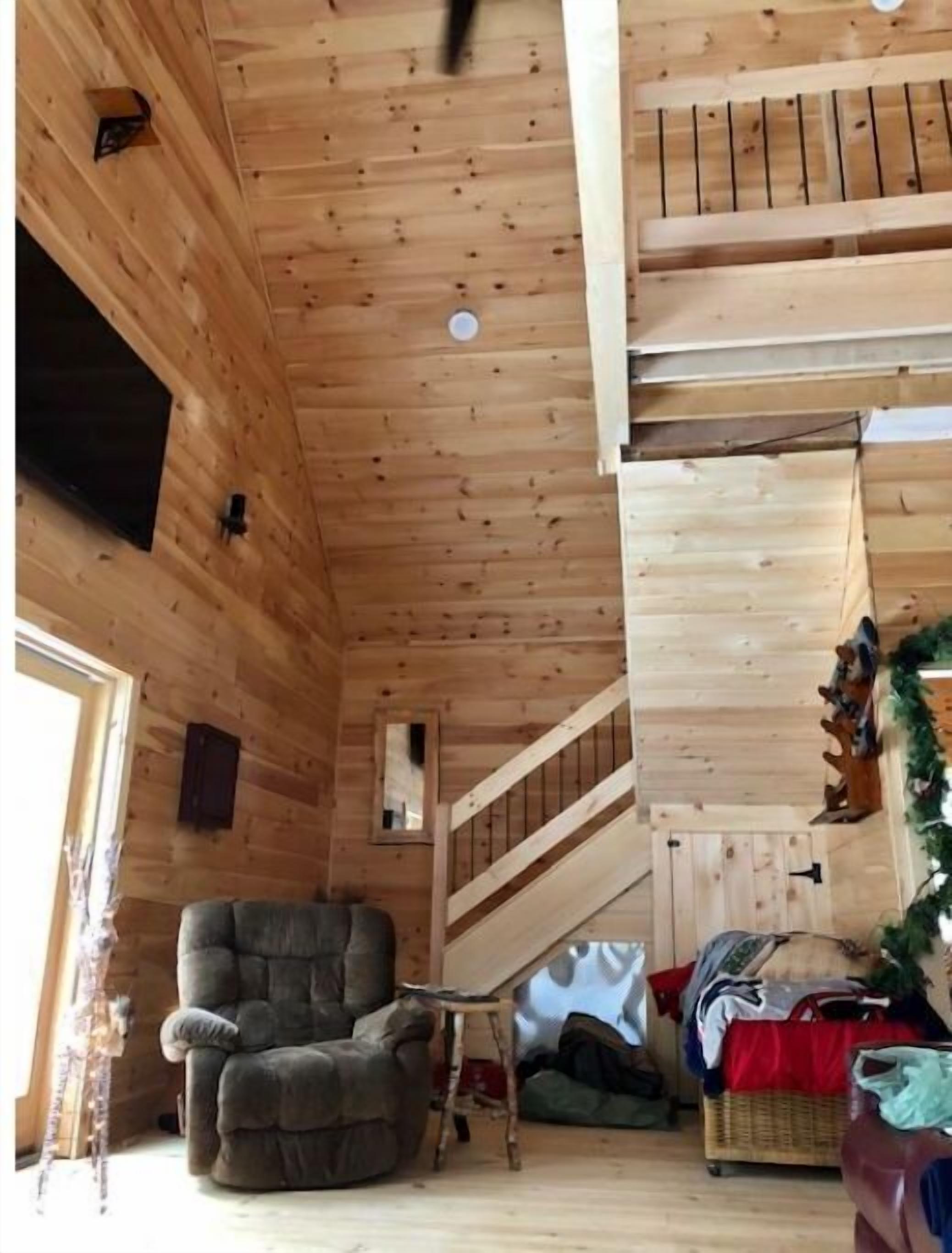 A wooden cabin interior with a cozy fireplace, a comfortable armchair, a bed with a red blanket, and a staircase leading to an upper level.
