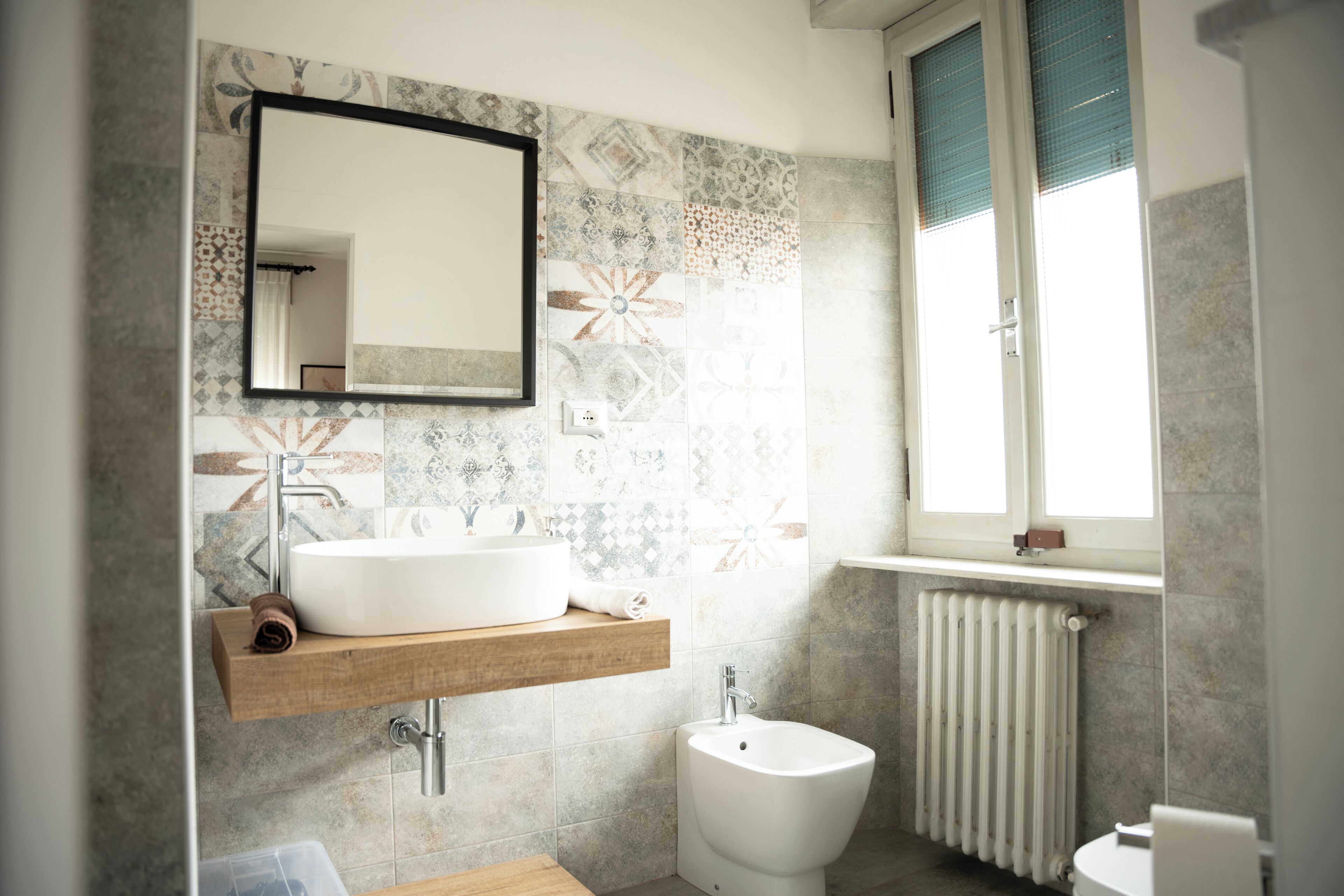 A modern bathroom with a large mirror, a white sink, a bidet, and a window with blinds.