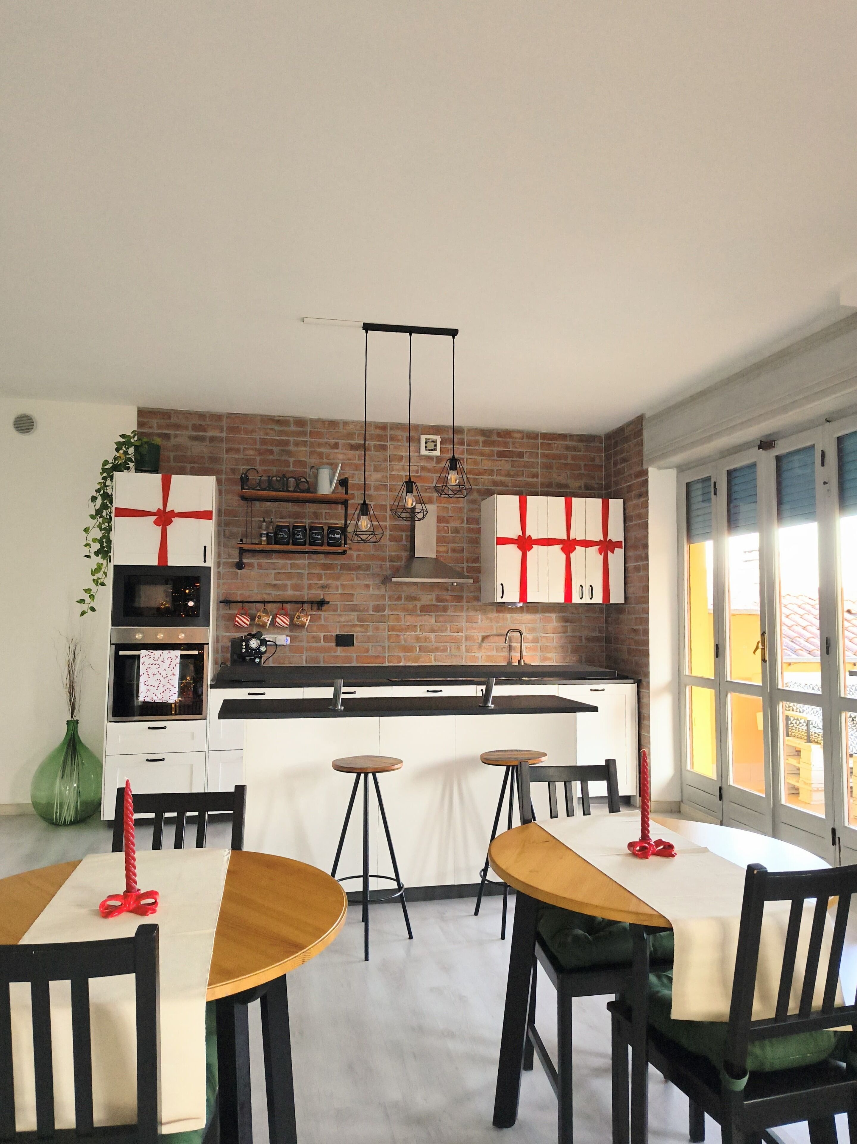 A modern kitchen with a brick wall, white cabinetry, and a central island. There are hanging pendant lights, a green plant, and a red gift box.
