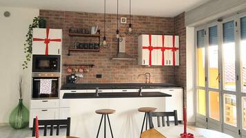 A modern kitchen with a brick wall, white cabinetry, and a central island. There are hanging pendant lights, a green plant, and a red gift box.