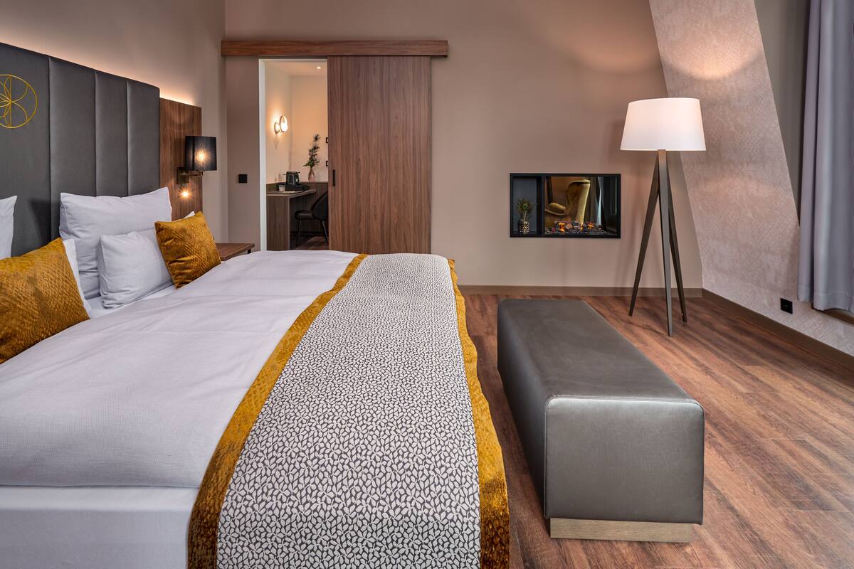 A hotel room with a large bed, a bedside table, a lamp, a bench, and a desk with a chair.