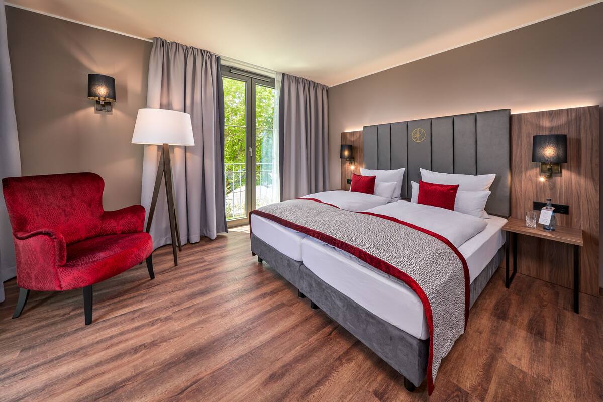 A hotel room with a large bed, a red armchair, a floor lamp, and a view of greenery through the window.