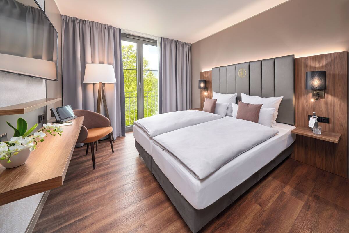A modern hotel room with a large bed, a desk with a chair, a lamp, and a view of greenery through the window.