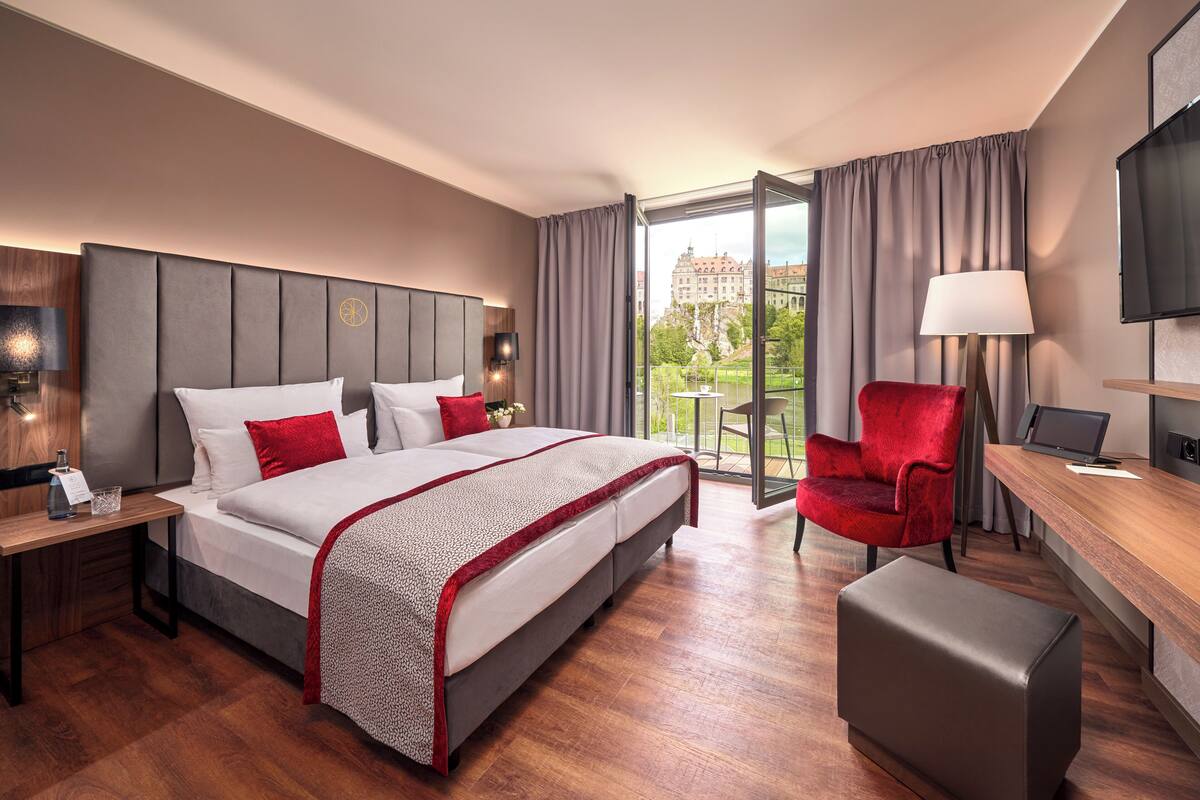 A hotel room with a large bed, a desk with a chair, a TV, and a balcony with a view of a building.