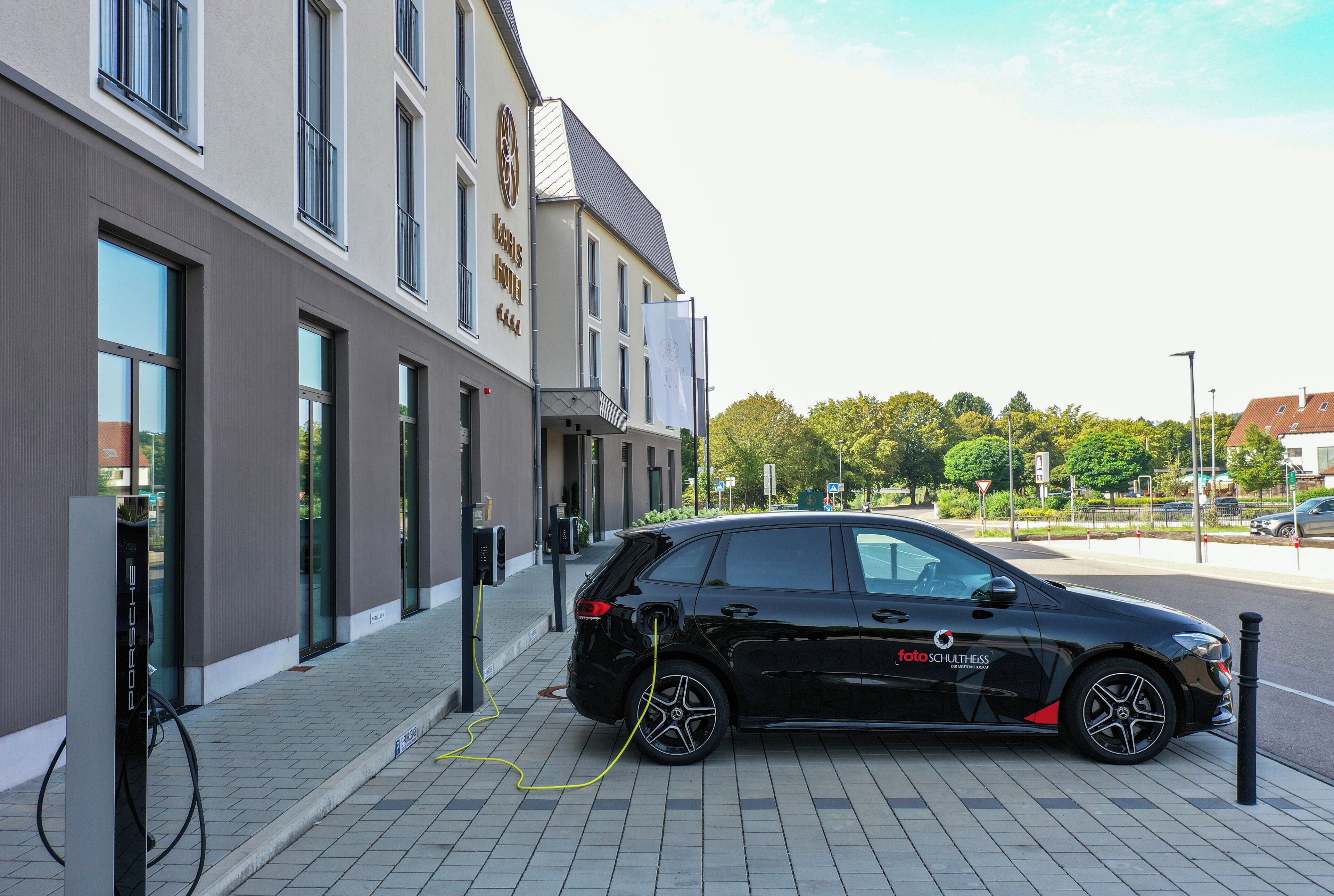 Une voiture noire est garée à une borne de recharge devant un immeuble doté de grandes fenêtres et d’une enseigne indiquant « Hôtel ».