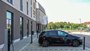 Une voiture noire est garée à une borne de recharge devant un immeuble doté de grandes fenêtres et d’une enseigne indiquant « Hôtel ».