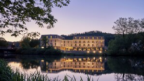 A multi-story hotel with a reflection in the water, surrounded by trees and a bridge. - Karls Hotel (Sigmaringen)