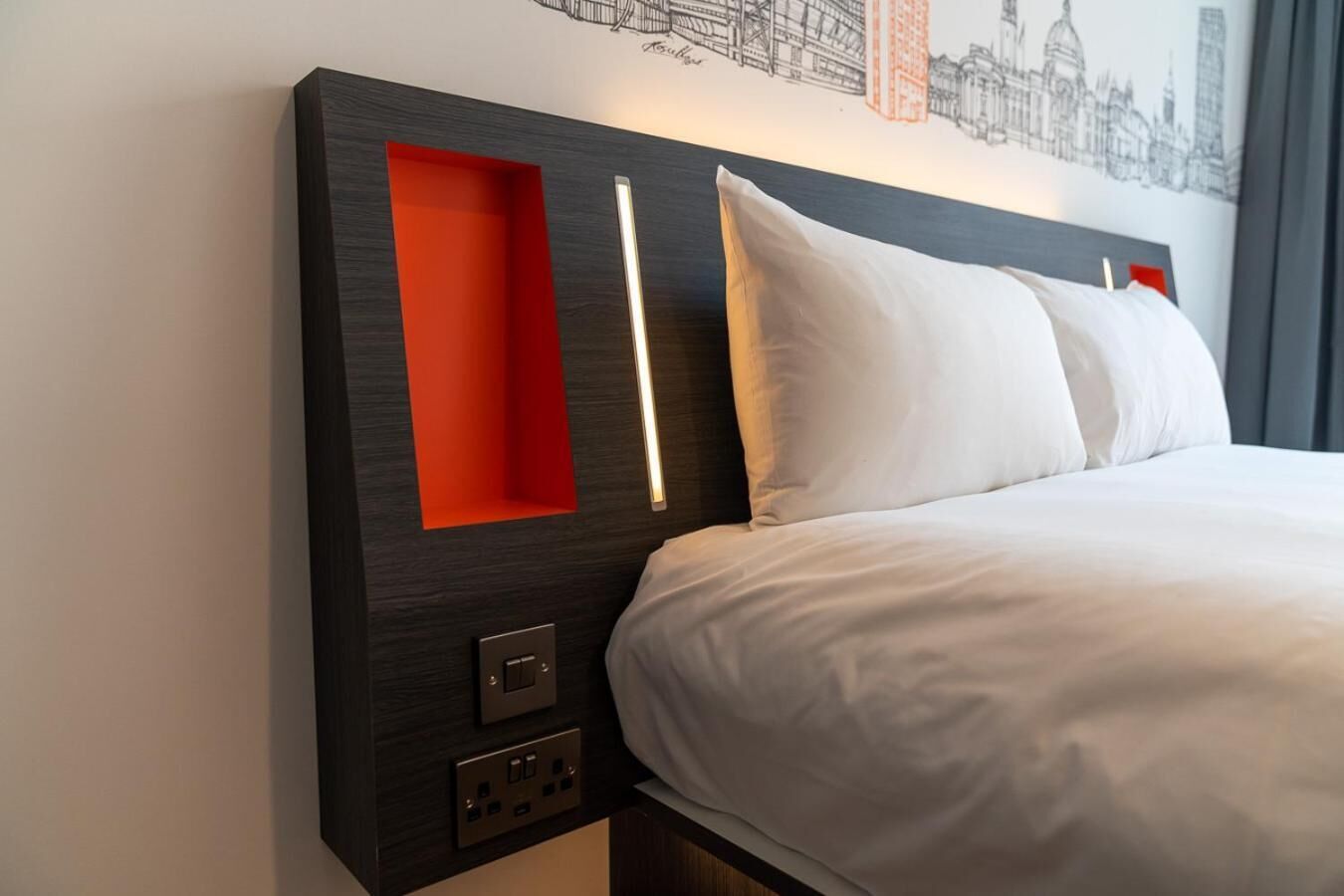 A modern hotel room with a large bed, a red accent wall, and a cityscape mural on the wall.