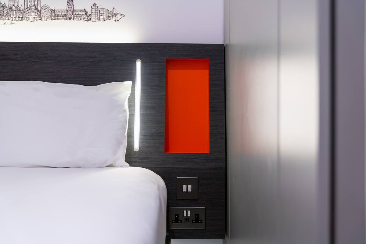 Et moderne hotelværelse med en pænt redt seng, et bybillede på væggen, et rødt farvefelt og en stikkontakt.