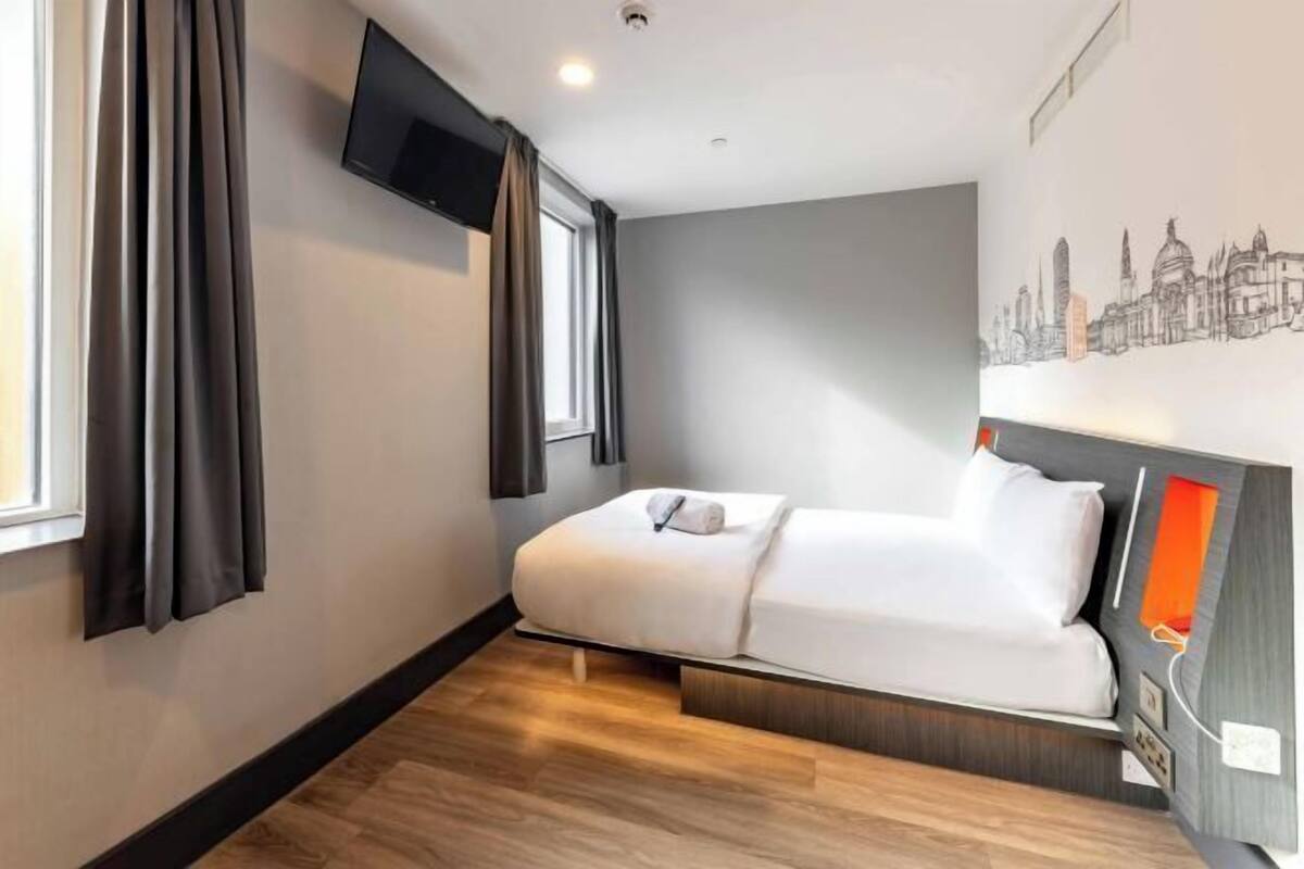 Et moderne hotelværelse med en seng, et fladskærms-tv ophængt på væggen og et bylandskabsmaleri.