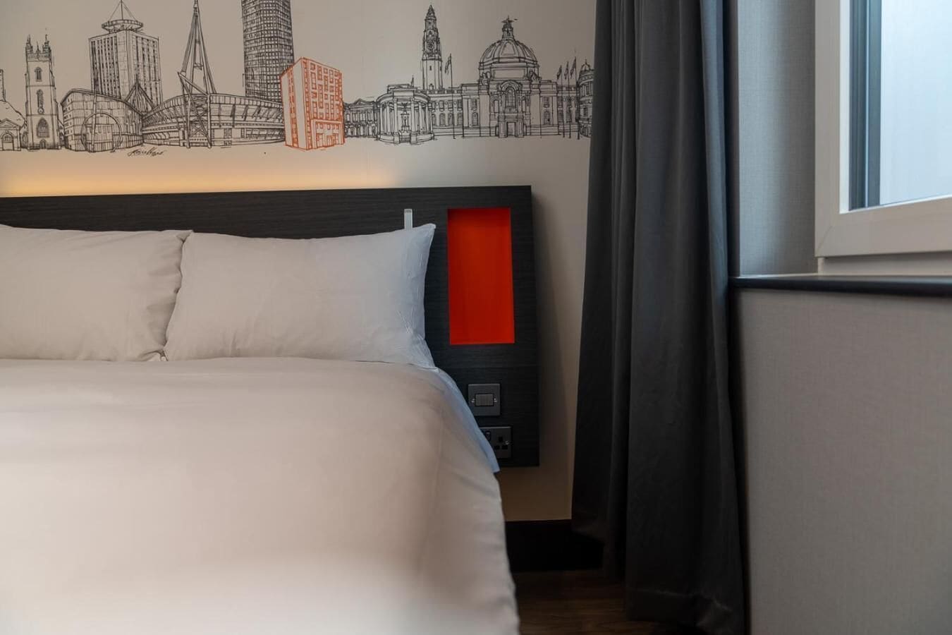 Una habitación de hotel con una cama, cabecero con detalle rojo y un mural en la pared con el horizonte de una ciudad.