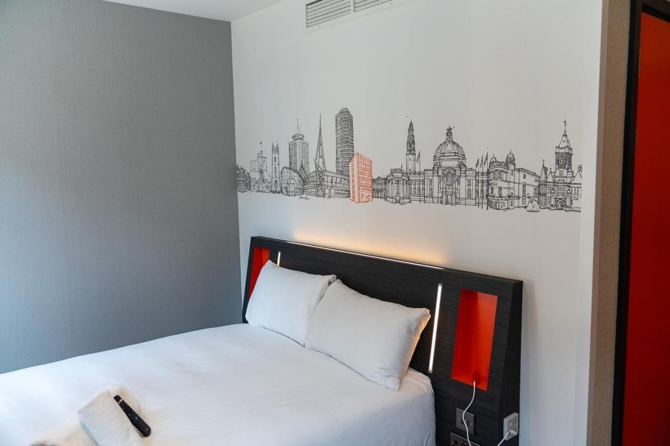 A hotel room with a bed, a cityscape wall art, and a red accent wall.
