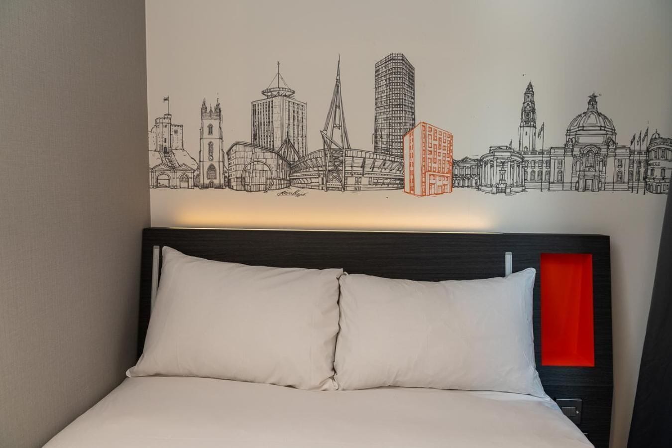 Una habitación de hotel con una cama, cabecera y un mural en la pared que muestra el horizonte de una ciudad.