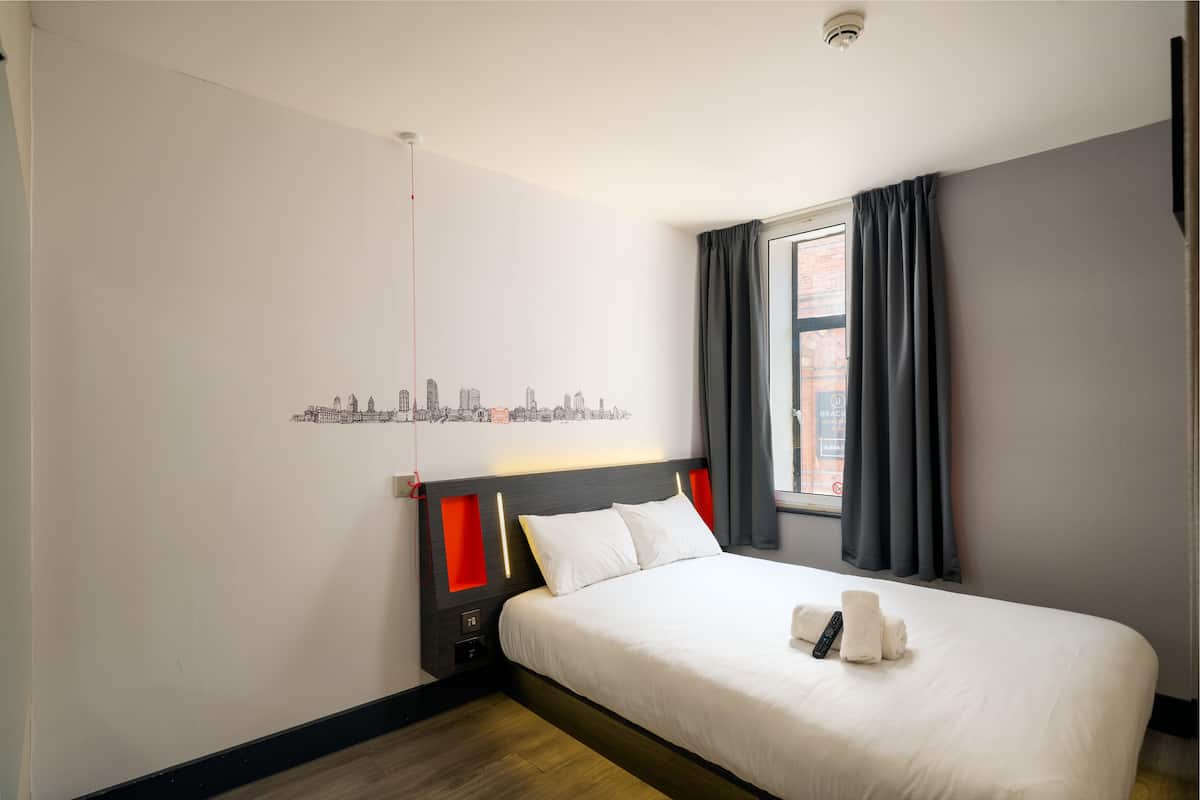 Et hotelværelse med en seng, bykunst på væggen, et vindue med gardiner og udsigt over byen.