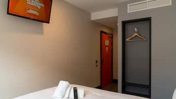 A hotel room with a bed, a remote control, a TV on the wall, a red door, and a wooden hanger on the wall.