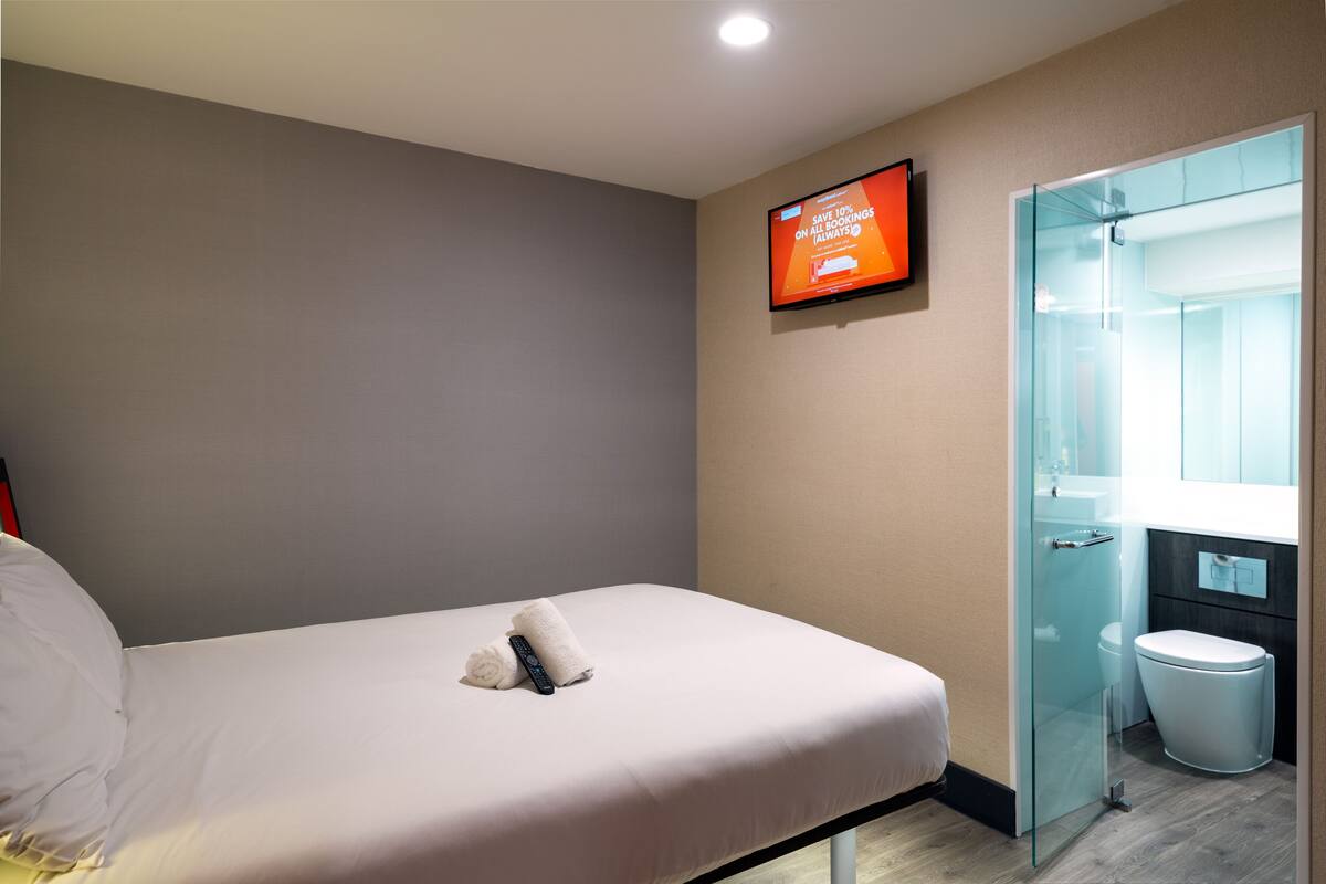 Et hotelværelse med en seng, et fjernsyn ophængt på væggen, en glas brusekabine og et hvidt toilet.