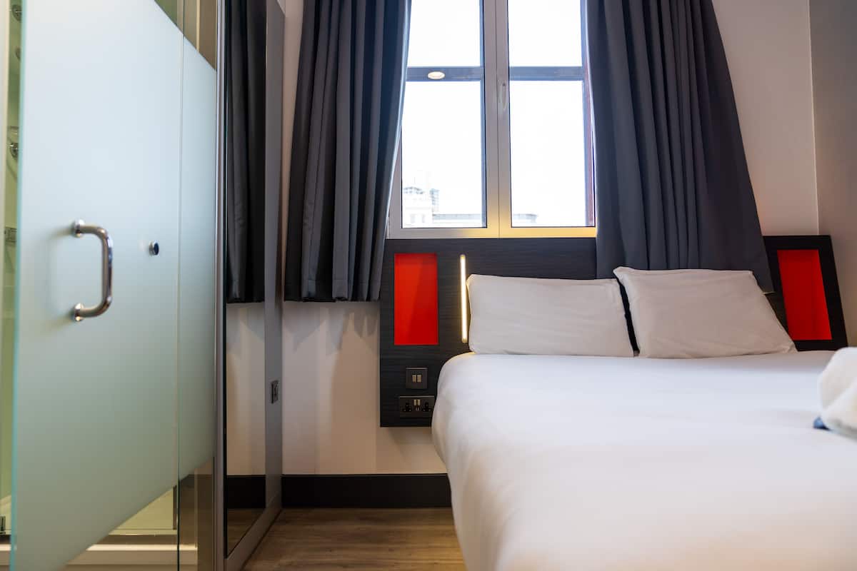 Et hotelværelse med en seng, et natbord, et vindue med gardiner og et panel monteret på væggen.