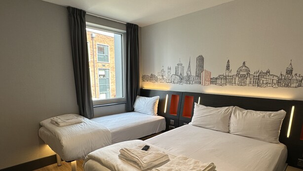 A hotel room with two beds, a cityscape mural on the wall, and a window with curtains. - easyHotel Cardiff (Cardiff)