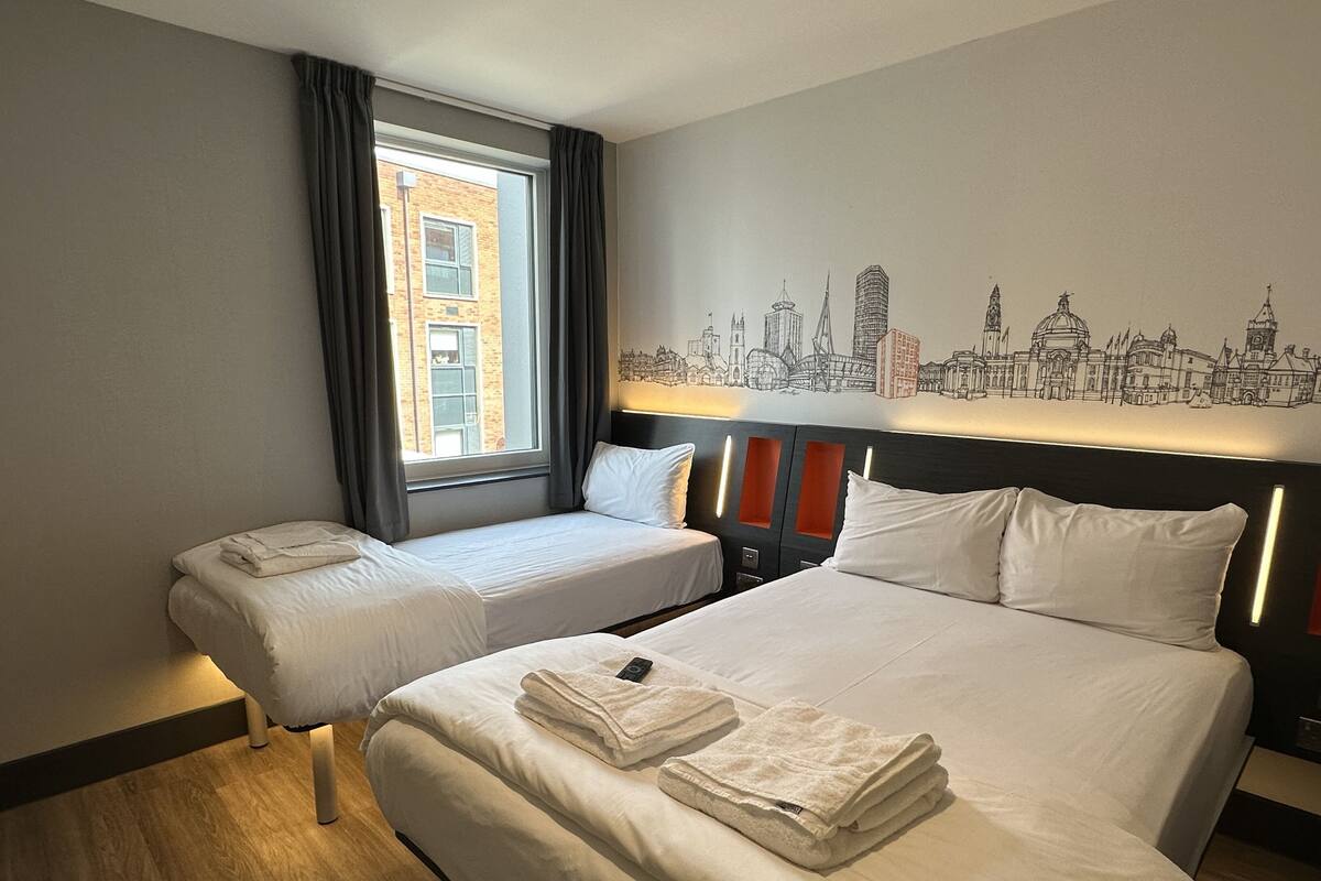 Et hotelværelse med to senge, et bybillede malet på væggen og et vindue med gardiner.