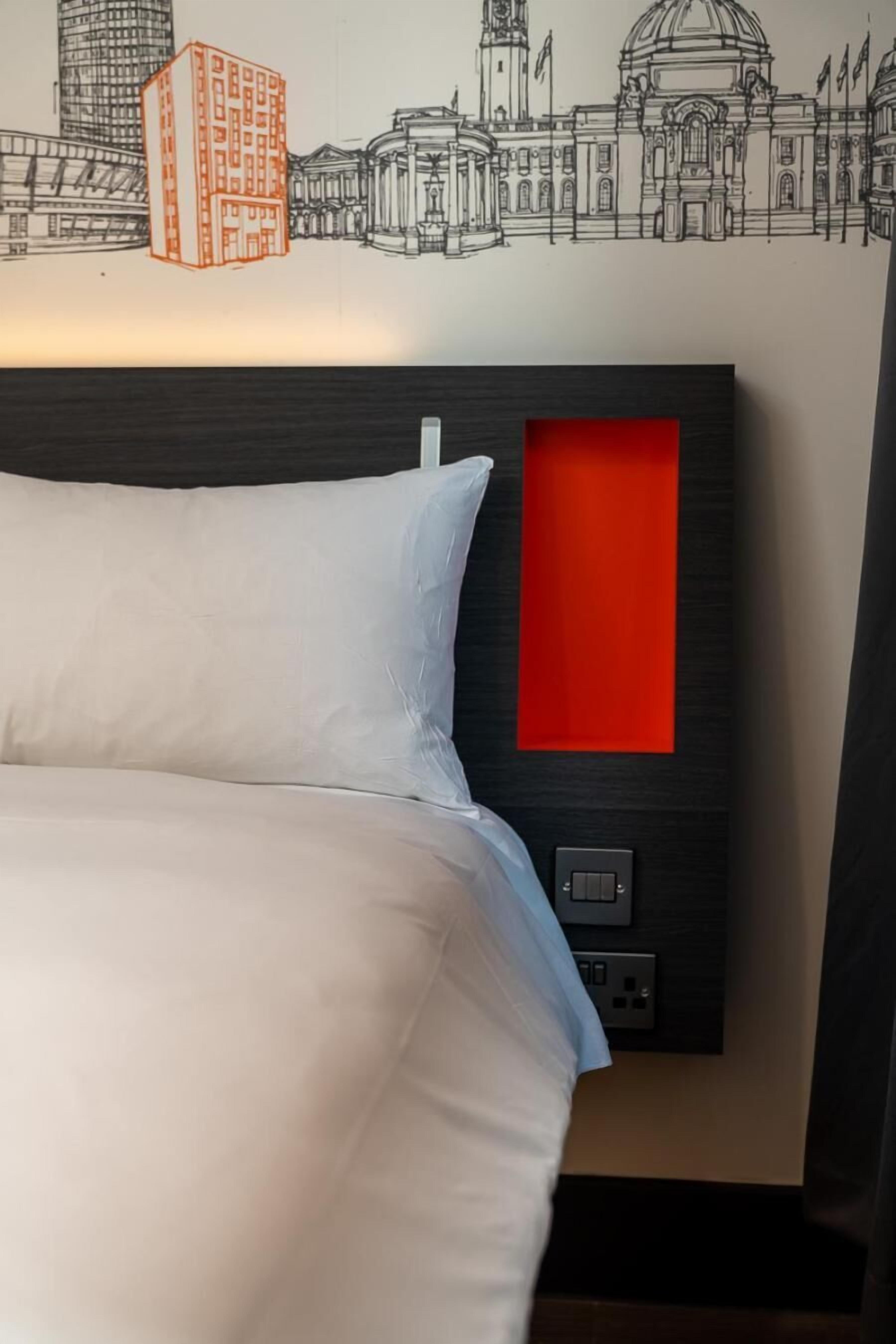 Une chambre d’hôtel avec un lit, une tête de lit ornée d’un panneau rouge et une fresque murale représentant un paysage urbain.