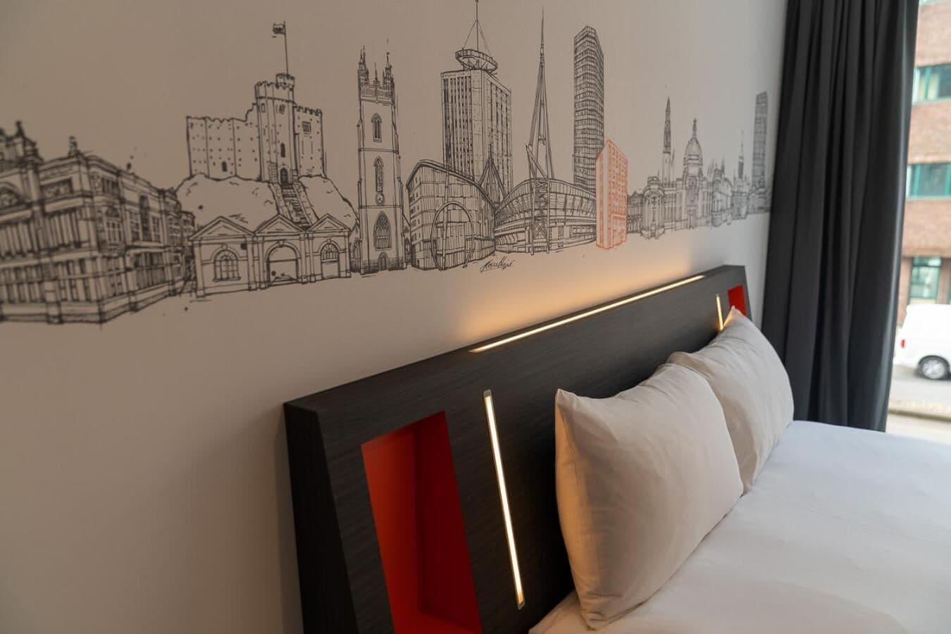 Une chambre d’hôtel avec un lit, une tête de lit agrémentée de touches de rouge et une fresque murale représentant un paysage urbain.