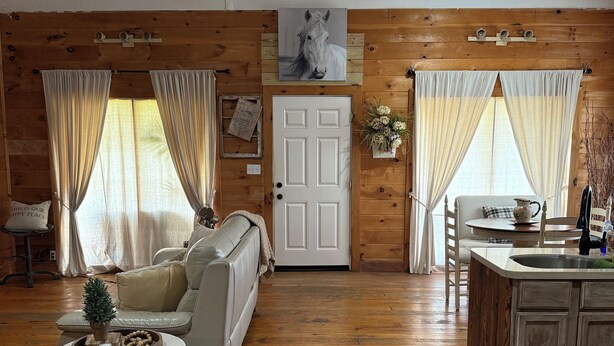 A cozy cabin interior with wooden walls, a white door, a sofa, a dining table, and a kitchen area. - Country Bliss (Tishomingo)