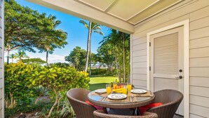 Une terrasse couverte, aménagée avec des meubles en osier, une table dressée avec des assiettes, des couverts et un panier de fruits.