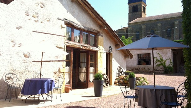 A rustic building with a stone facade, a wooden door, and a blue awning. There are outdoor tables and chairs, and a stone wall in the background. - La Grange : La Chambre Vendange (Coutouvre)
