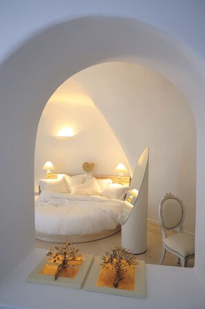 Ein Schlafzimmer mit gewölbter Decke, einem Bett mit weißen Leinen, zwei Nachttischlampen, einem runden Spiegel und einem Sessel.
