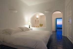 A bedroom with a bed, a nightstand, a lamp, and a view of the sea through an open door. - White (Santorini)