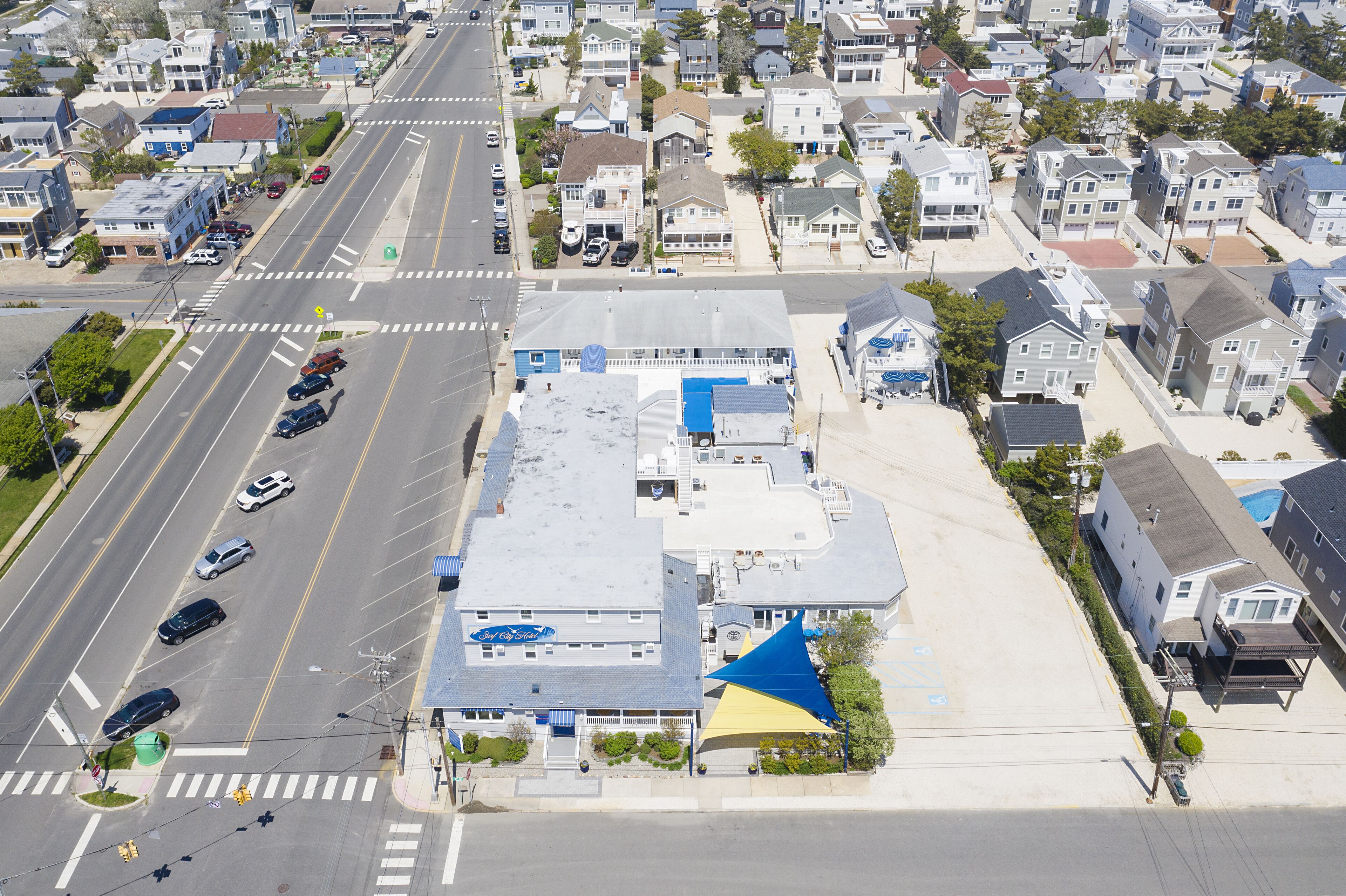 Vue aérienne d’un quartier résidentiel avec un hôtel reconnaissable à son auvent bleu et jaune, un parking et une route où circulent des voitures.