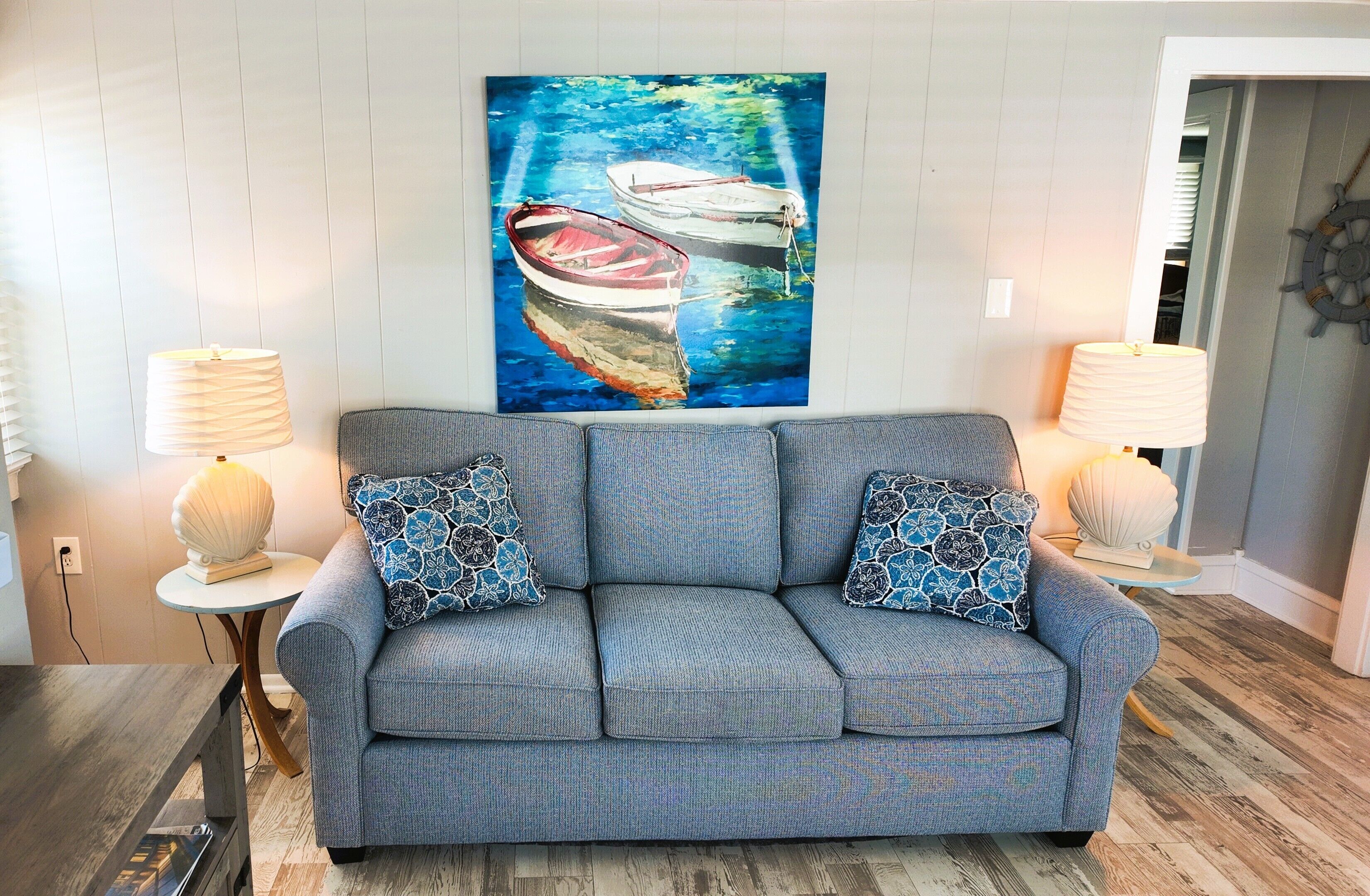 A living room with a grey sofa, two patterned throw pillows, a painting of boats on water, and two lamps on side tables.