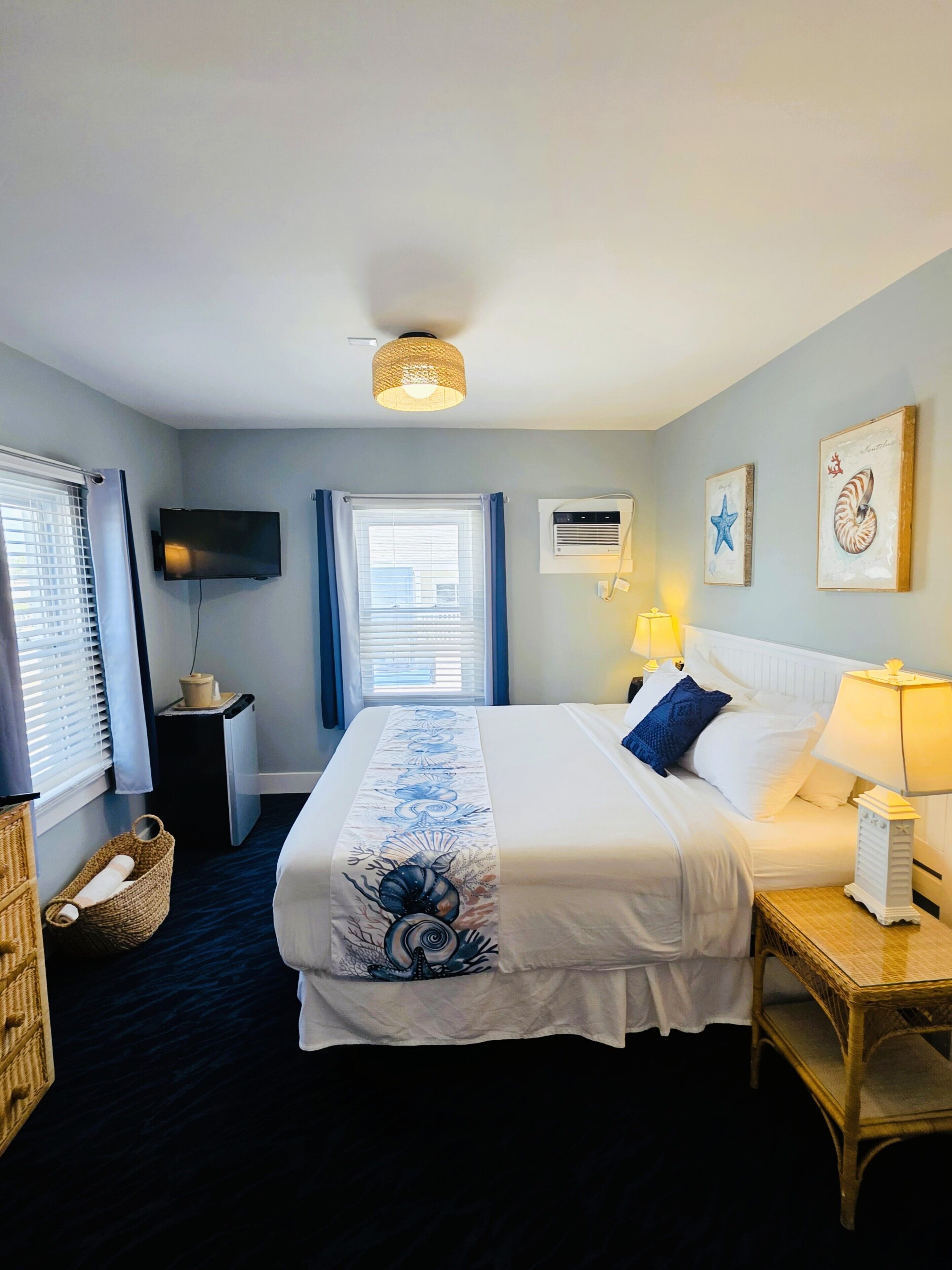 A hotel room with a bed, a television, a window, a wall-mounted air conditioner, and a lamp.