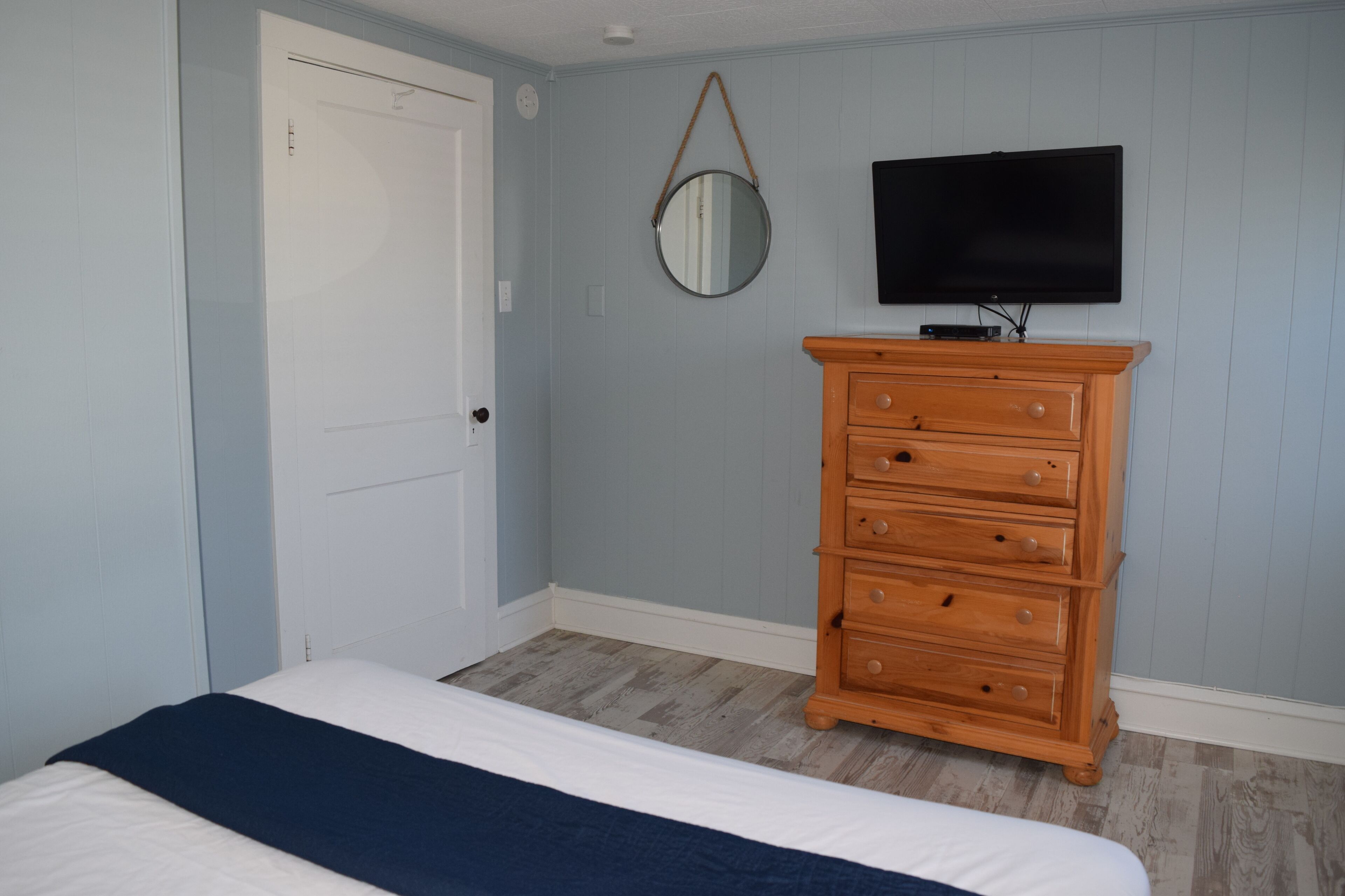 Une chambre à coucher avec un lit, une commode en bois, un miroir et un téléviseur fixé au mur.