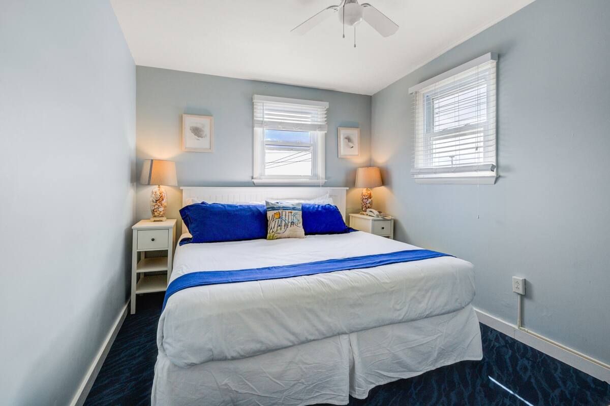 A bedroom with a large bed, two bedside tables with lamps, a ceiling fan, and two windows with blinds.