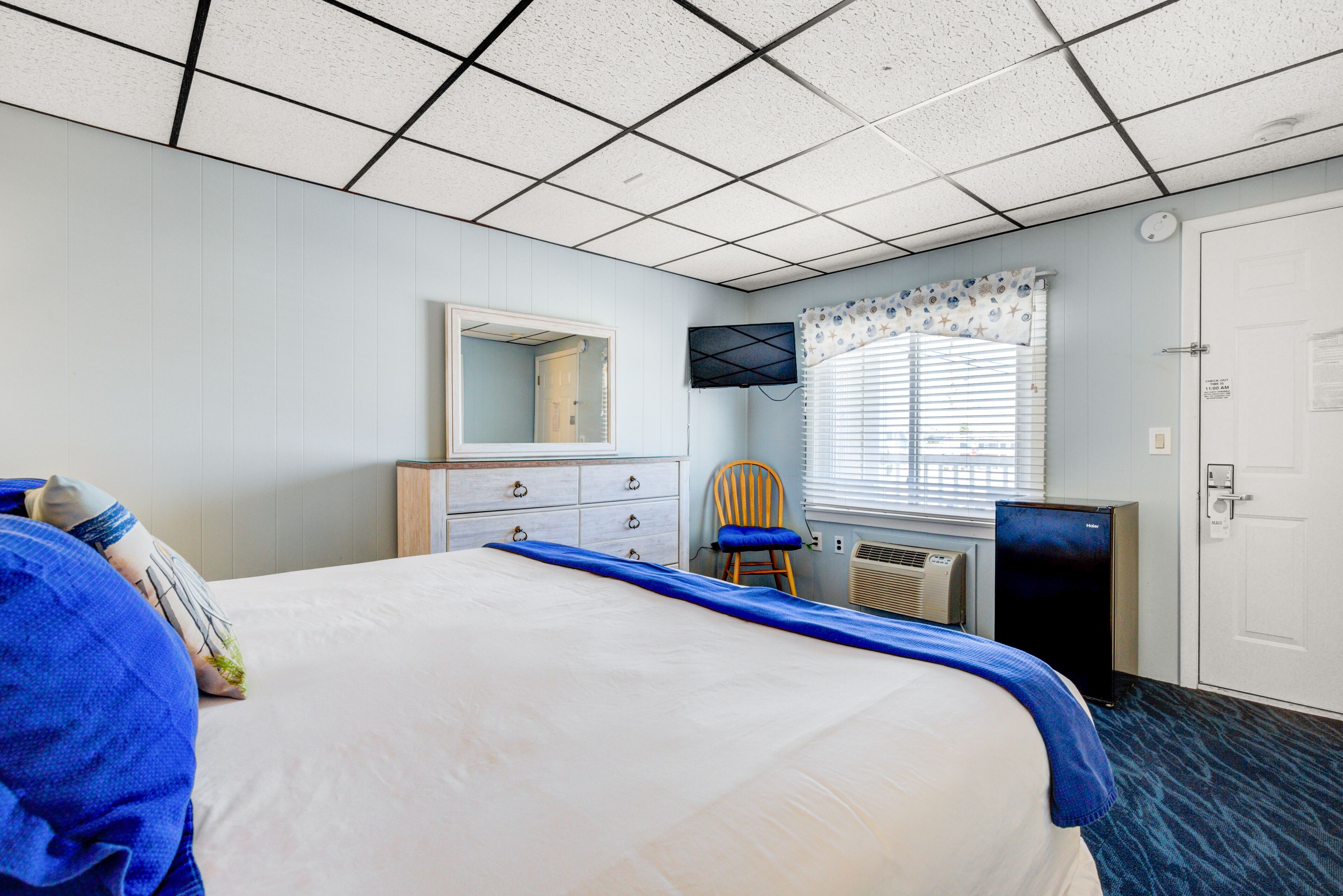 A hotel room with a large bed, a dresser, a chair, a television, and a window with blinds.