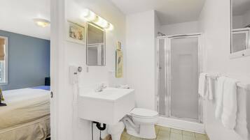 Une salle de bain avec un lavabo, des toilettes et une douche, le tout de couleur blanche. Un miroir avec des lumières est placé au-dessus du lavabo, et un porte-serviettes est fixé au mur.