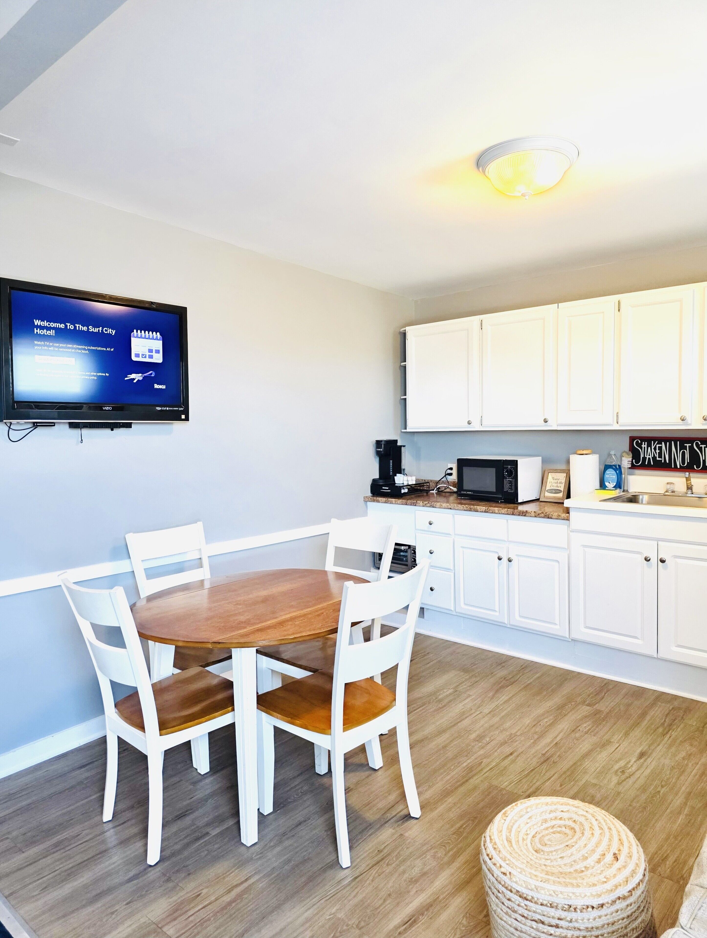 A room with a dining table, chairs, a television mounted on the wall, and a kitchen area with cabinets and appliances.